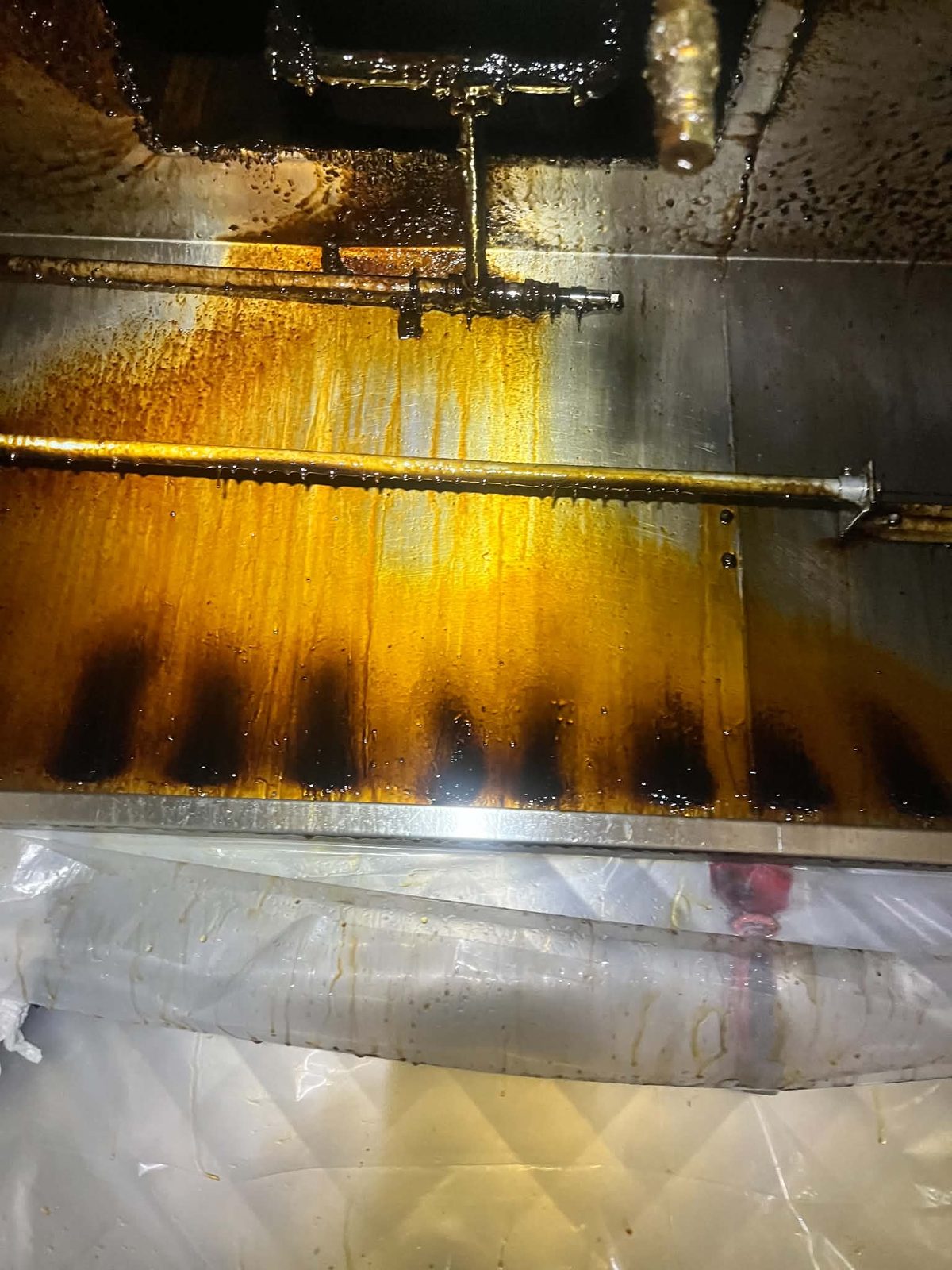 Amber degreaser running down the inside of a kitchen exhaust plenum, dissolving black grease deposits, with plastic containment sheeting spread underneath to catch the runoff